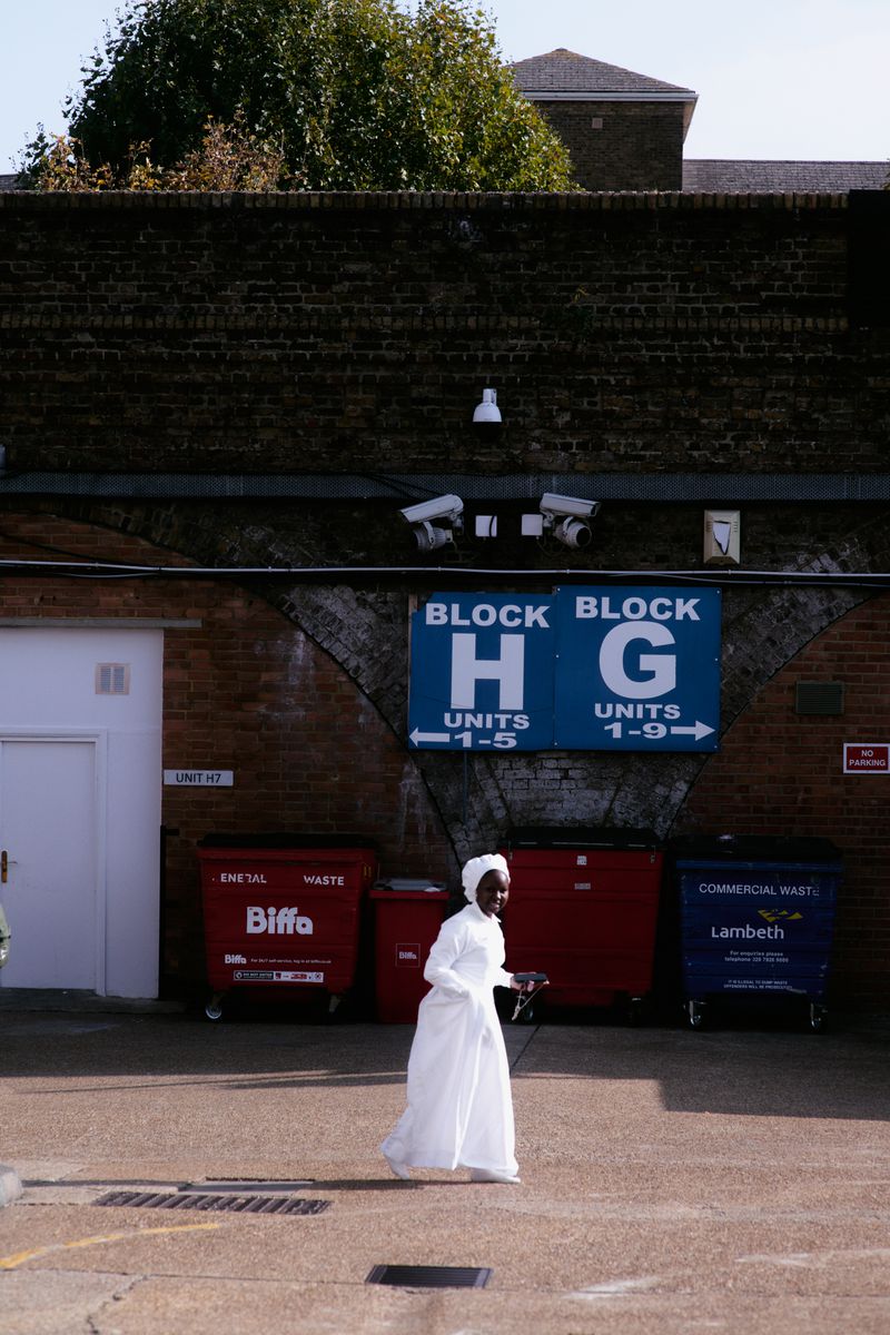 Model in a flowing white gown and headwrap walking past industrial brick arches and waste bins in direct sunlight.