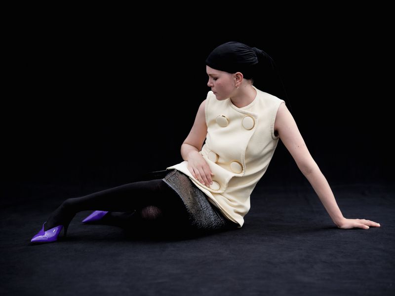 Model seated on a black seamless background wearing a cream double-breasted top and purple heels