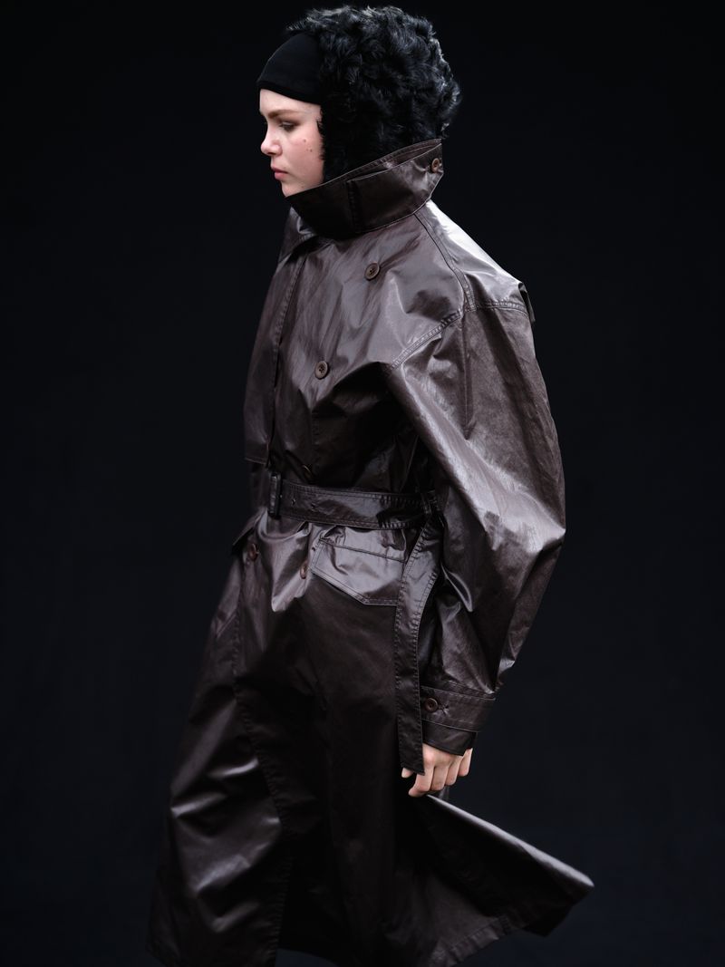 Model wearing a dark glossy coat with a high collar and matching headpiece against a plain dark background.