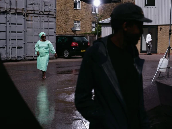 Woman in a mint green dress and matching headwrap walking across wet pavement with a silhouetted man in the foreground.