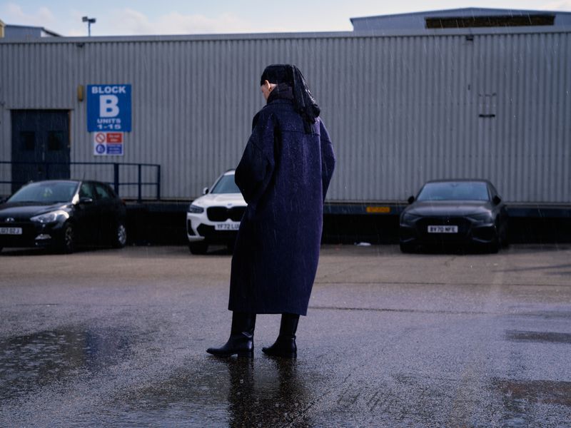 Model Thea Almqvist in a long navy coat and black boots standing in a wet parking lot outside an industrial building.