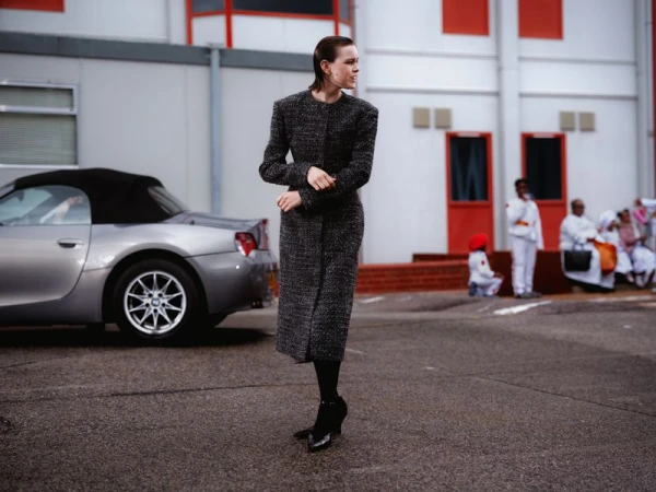 Model Thea Almqvist in a tailored charcoal tweed coat walking across a paved lot beside a silver convertible sports car.
