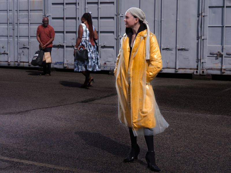 Model Thea Almqvist in a bright yellow coat with translucent overlay and headscarf posing in front of grey shipping containers.