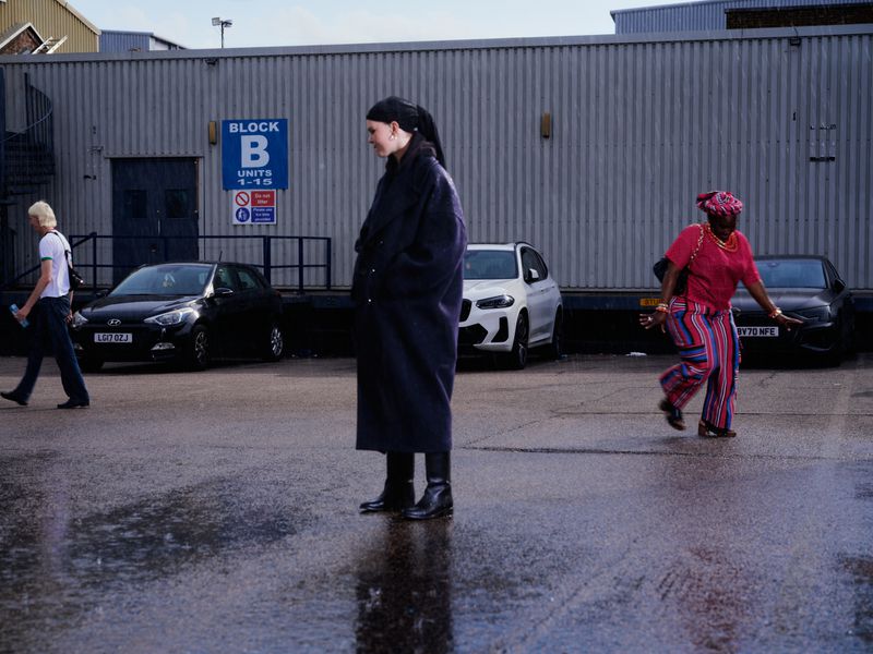 Model Thea Almqvist in a long dark navy coat standing on wet asphalt in a parking lot with figures moving in the background.