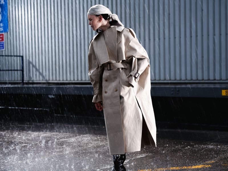 Model wearing an oversized beige belted trench coat and headscarf in the rain against a corrugated metal industrial wall.