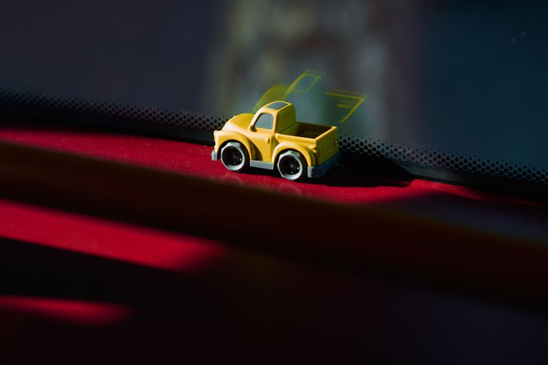 A miniature yellow toy pickup truck resting on a red glossy surface next to a car windshield with a dot matrix border.