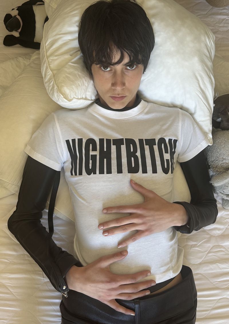 Brunette laying on a white bed wearing a t-shirt with 'NIGHTBITCH' printed on it, a black undershirt, and black pants.