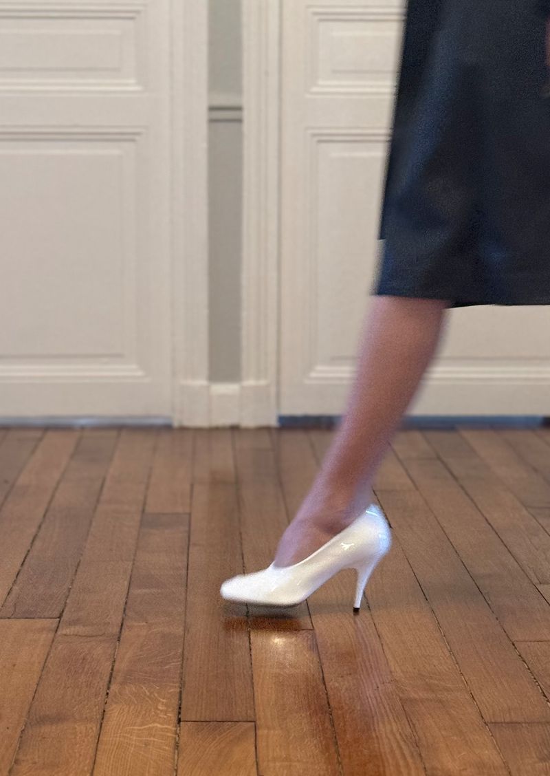 Close-up of a woman's leg and white patent leather stiletto heel on a wooden floor, with blurred motion, and white doors in the background.