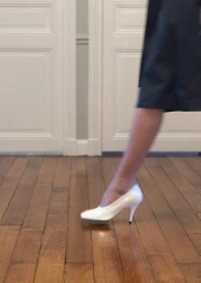 Model walking across a wooden parquet floor wearing a glossy white high-heeled pump and a dark skirt.