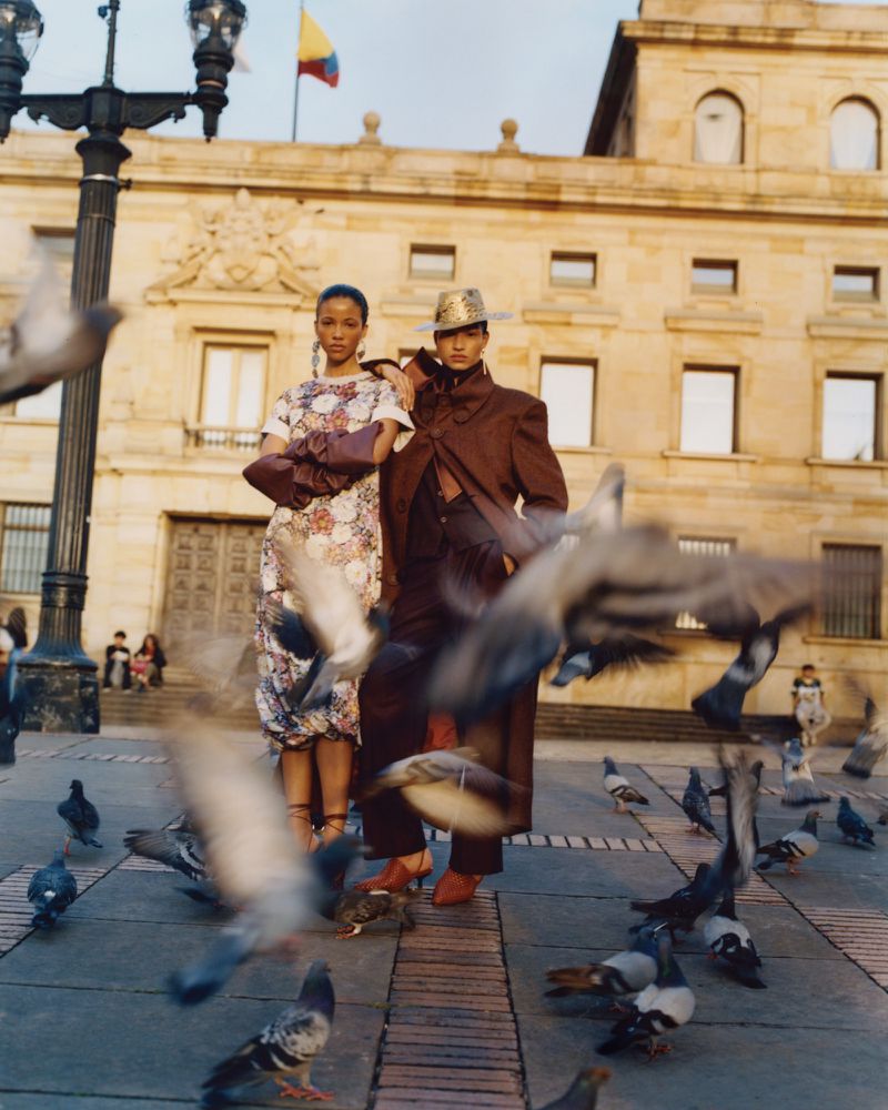 Two fashion models posing in a historic public square with pigeons flying around them in front of a grand stone building.