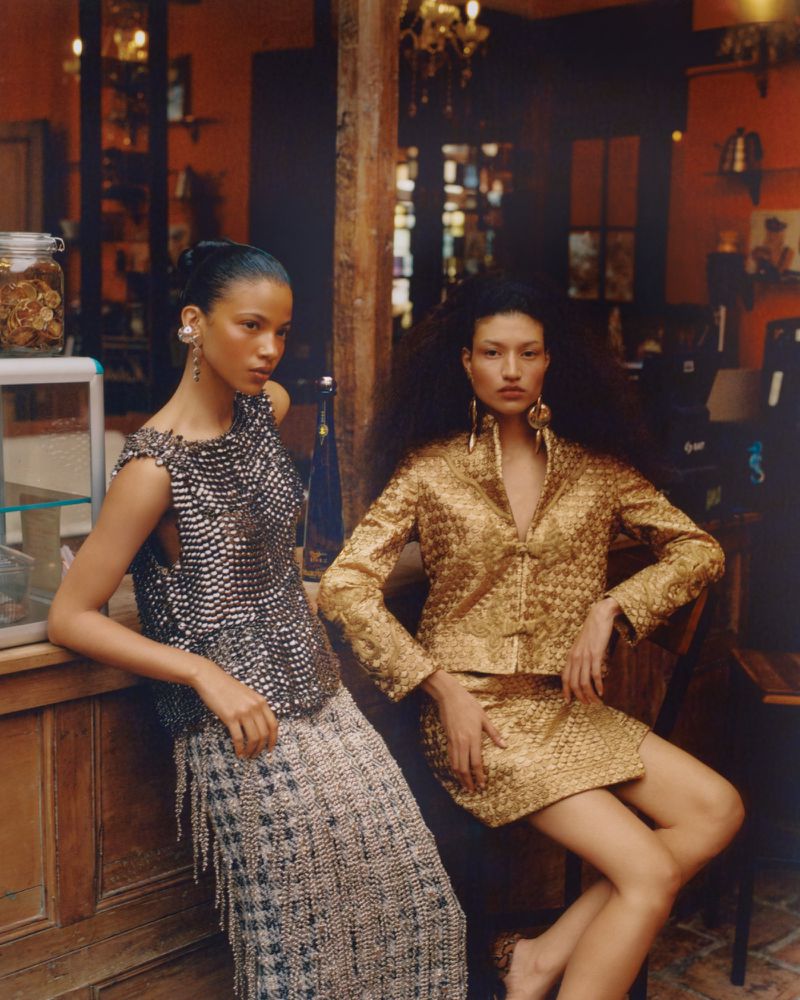 Two models posing inside a warm-lit rustic bar wearing textured metallic and gold designer outfits for Vogue Latin America.