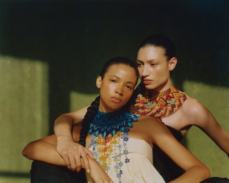 Two women wearing ornate beaded statement necklaces and strapless tops posing together in direct sunlight against an olive wall.