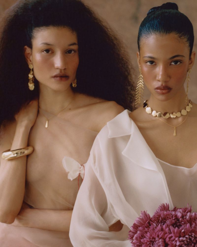 Honey Ordonez and Keny Salgado wearing gold jewelry and sheer white apparel against a warm neutral backdrop.