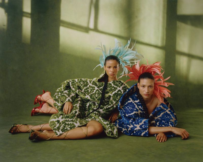 Two models reclined on a green studio floor wearing ornate patterned coats, strappy heels, and sculptural feather headpieces.