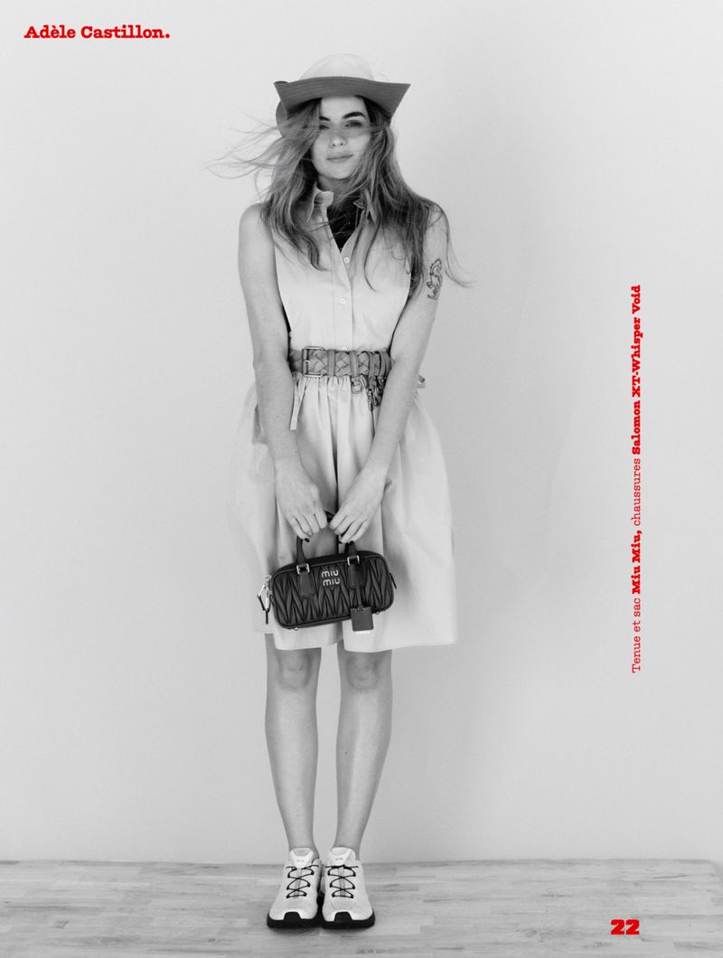 Adèle Castillon wearing a sleeveless dress, wide belt, and hat while holding a small black quilted handbag in a studio portrait.