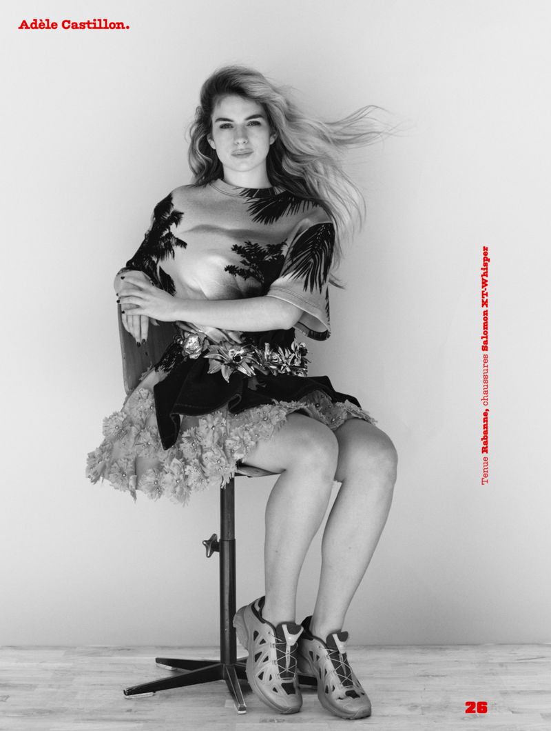 Black and white portrait of Adele Castillon seated on a studio stool wearing a patterned top and sneakers against a plain background.