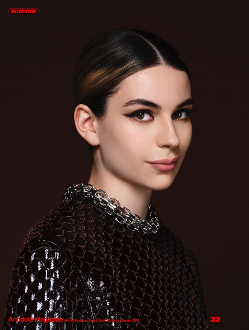 Portrait of a woman with winged eyeliner and a slicked hairstyle wearing a textured dark top and silver chain collar.