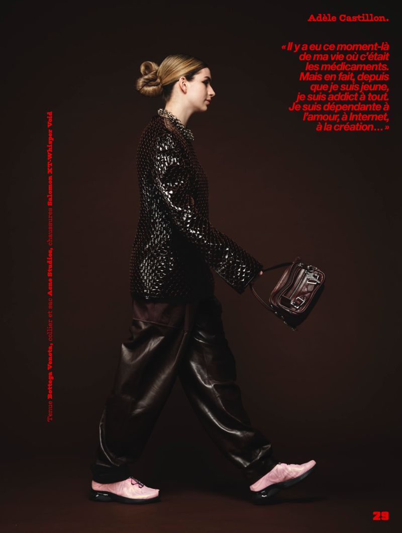 French artist Adele Castillon walking in profile wearing a black textured jacket, dark wide-leg trousers, and pink sneakers.