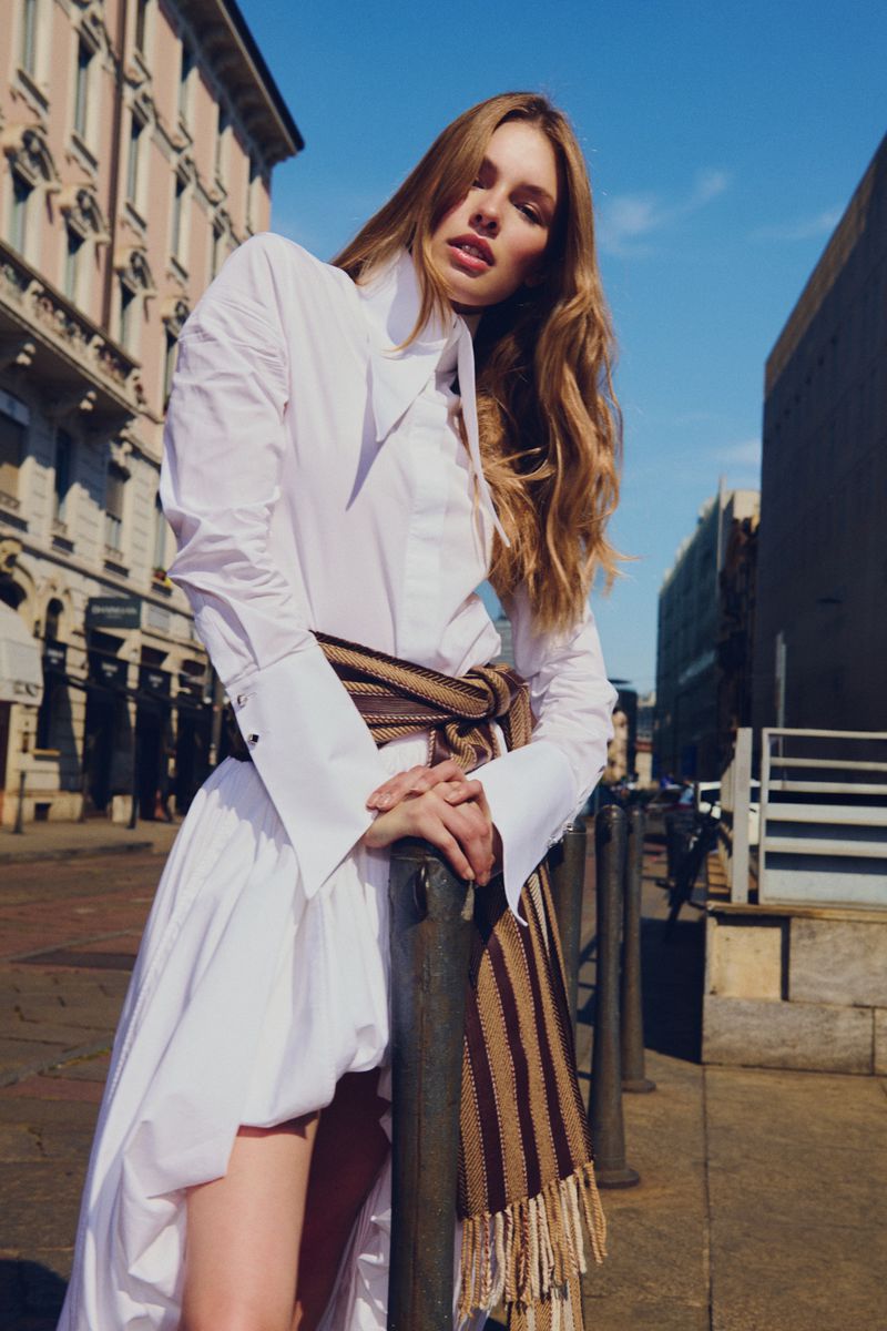 Model posing in an oversized white shirt dress with a striped fringed scarf tied at the waist on a city street.