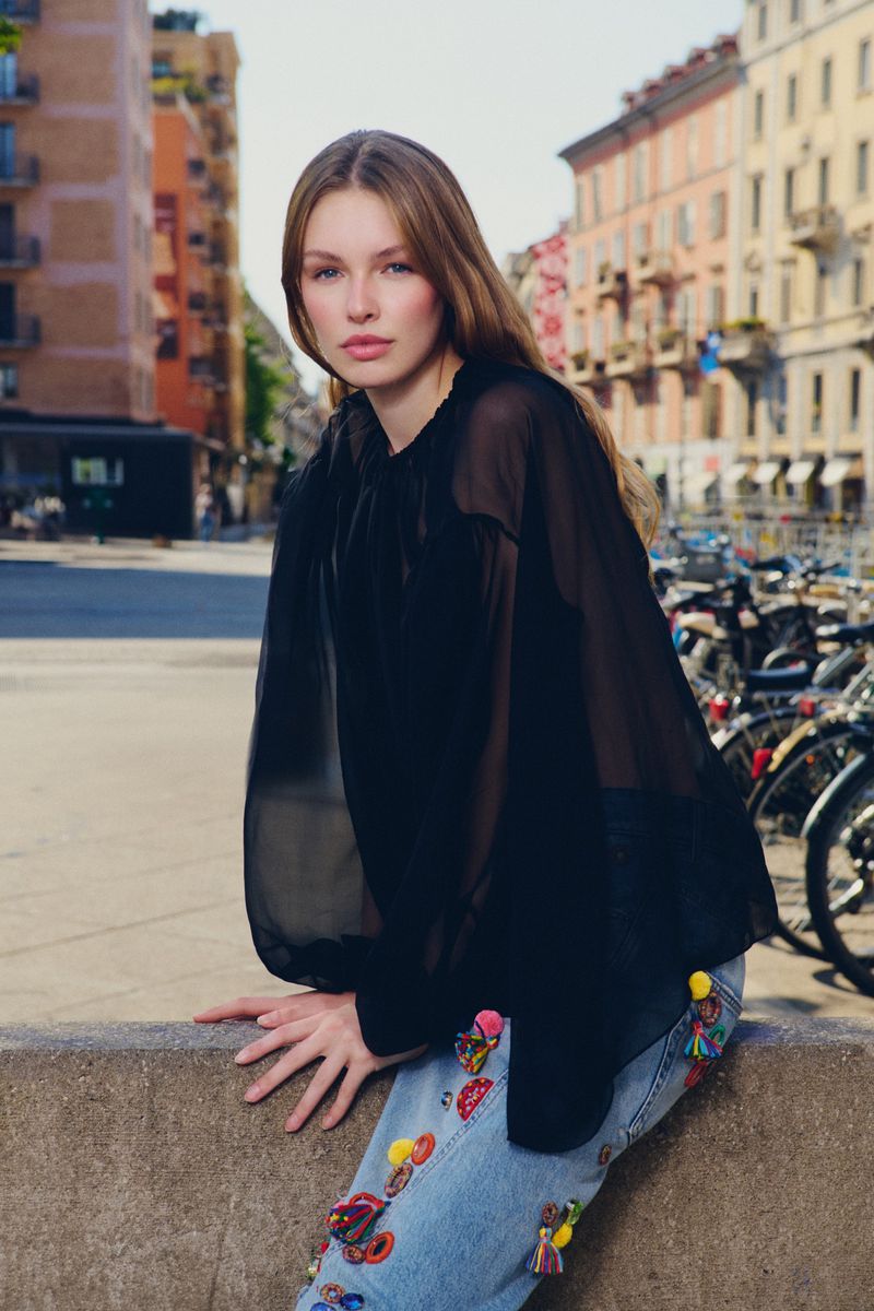 Model wearing a sheer black blouse and embellished denim jeans posing outdoors on a concrete ledge in a city square.
