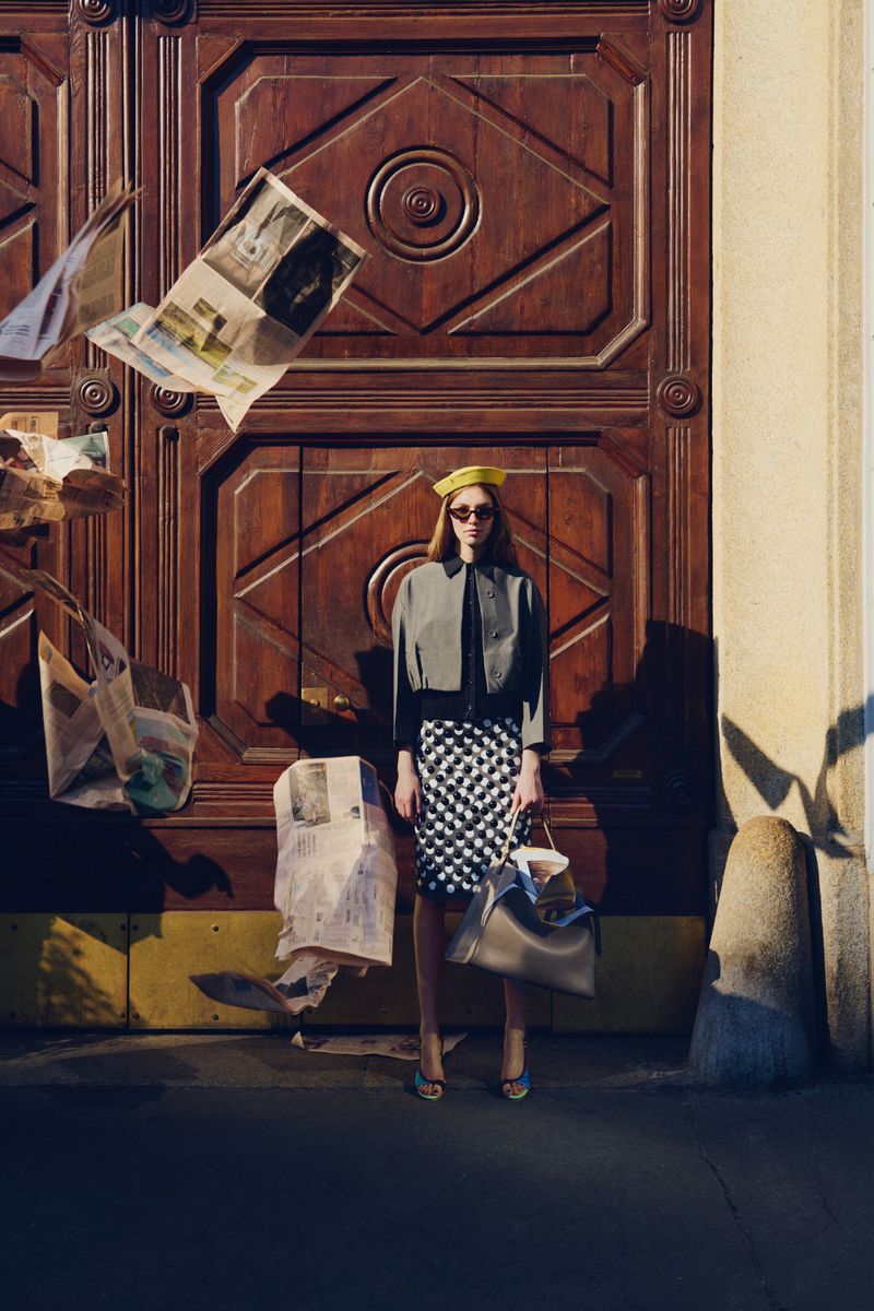 Fashion model wearing a yellow hat, grey cropped jacket, and patterned skirt standing by a wooden door with flying newspapers.