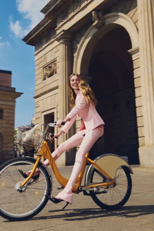 Woman in a tailored blush pink pantsuit riding a yellow bicycle in front of a classical stone archway in sunny daylight.