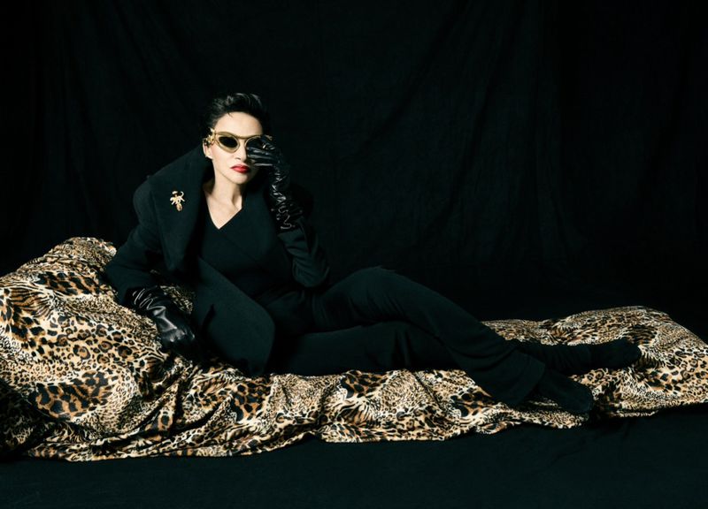 Farida Khelfa in an all-black suit, leather gloves, and gold sunglasses reclining on leopard-print fabric against a black studio backdrop.