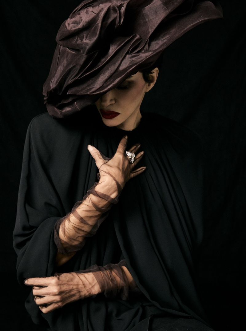 Model Farida Khelfa wearing an oversized brown sculptural hat, sheer ruched gloves, and black draped cape against a dark background.