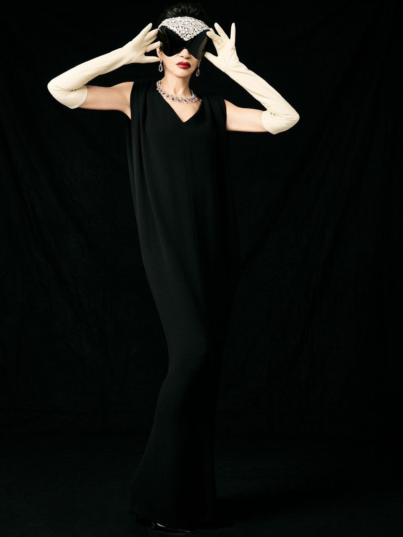Farida Khelfa posing in a sleeveless black column gown, cream opera gloves, and crystal-embellished glasses against a black backdrop.