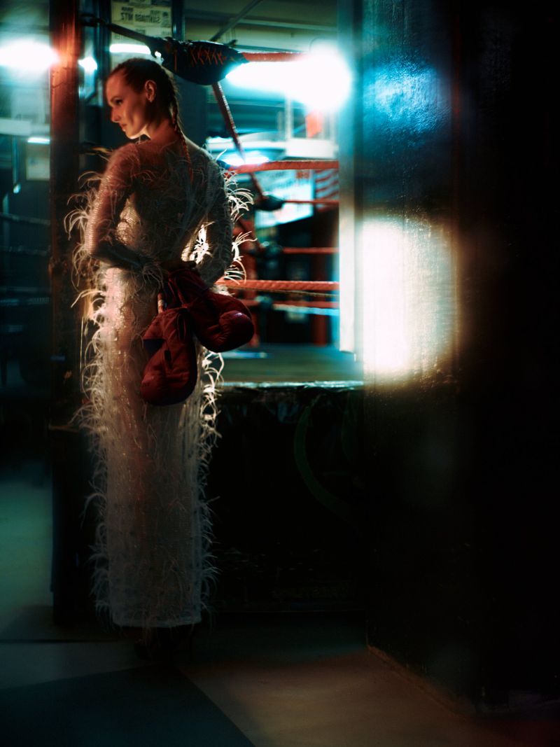 Model Anastasiia Koval in a white feathered gown holding red boxing gloves beside a boxing ring.