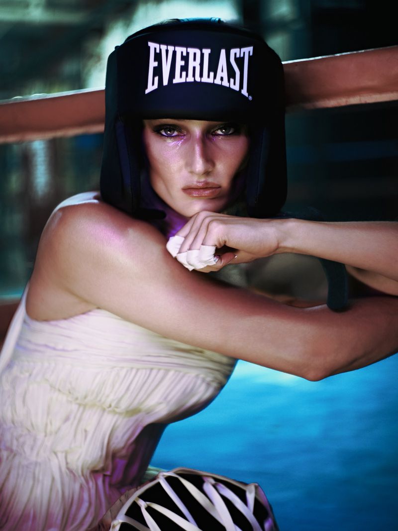 Model Anastasiia Koval wearing a black Everlast boxing headgear and off-white pleated dress while leaning on a boxing ring rope.