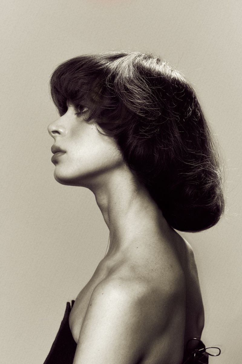 Model in profile showcasing a voluminous rounded dark bob haircut with bare shoulders against a neutral studio backdrop.