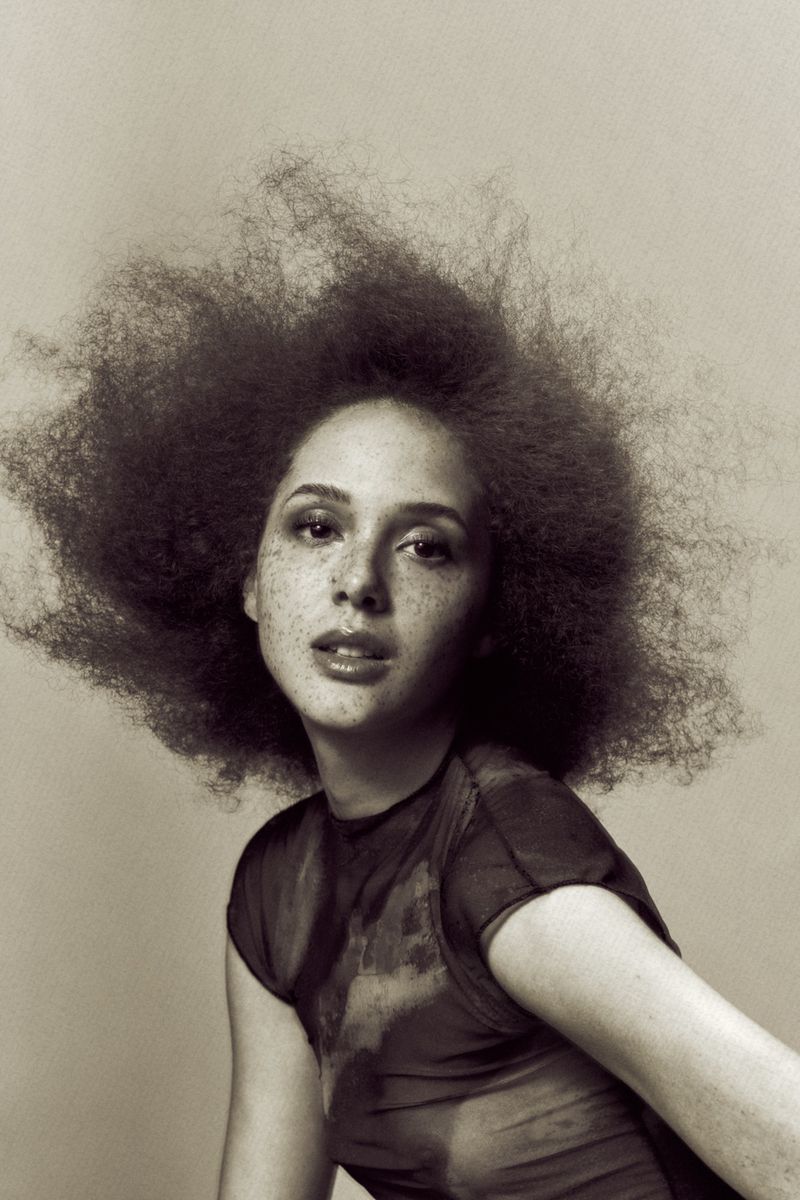Model Polina Dvoretskaia with voluminous textured hair and freckles posing in a dark top against a neutral studio backdrop.