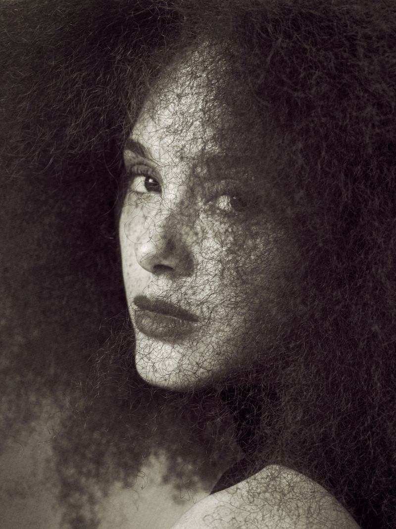 Close-up portrait of a woman with voluminous textured hair casting fine shadows across her freckled face in monochrome.