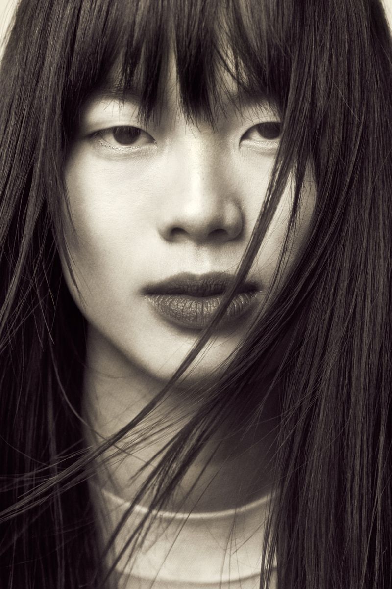 Close-up portrait of an East Asian woman with straight dark bangs and loose hair strands falling across her face.