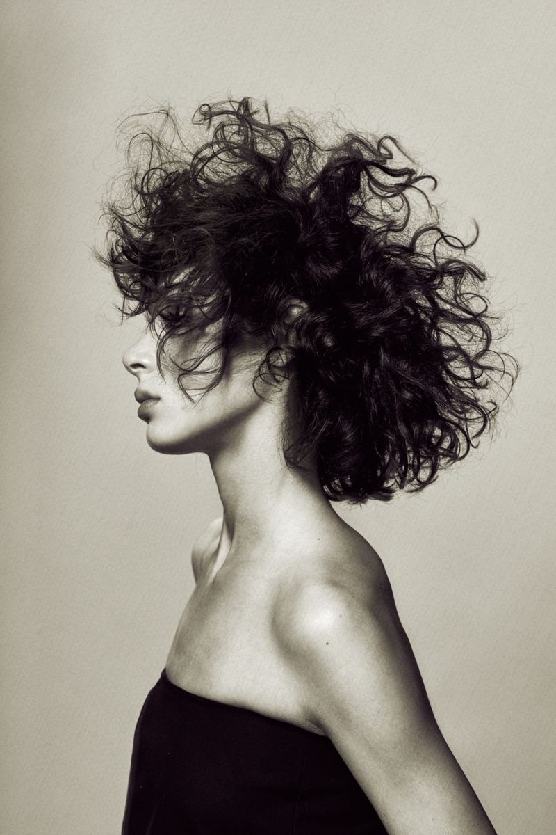 Profile portrait of a woman with voluminous textured curly hair wearing a strapless black top against a studio background.