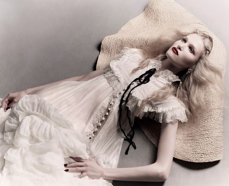 Model Sascha Raj lying on a grey studio floor resting her head on an oversized straw hat while wearing a ruffled white lace dress.