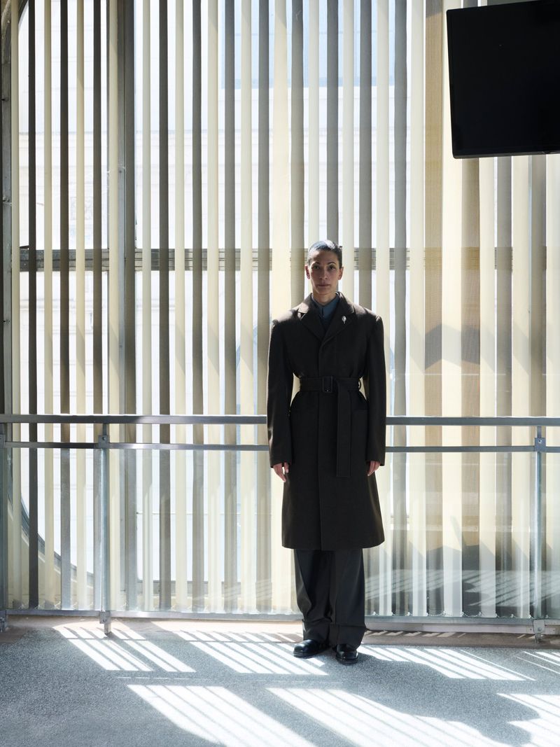 Model wearing a dark tailored belted overcoat and wide trousers standing in front of tall vertical window blinds casting slatted shadows.