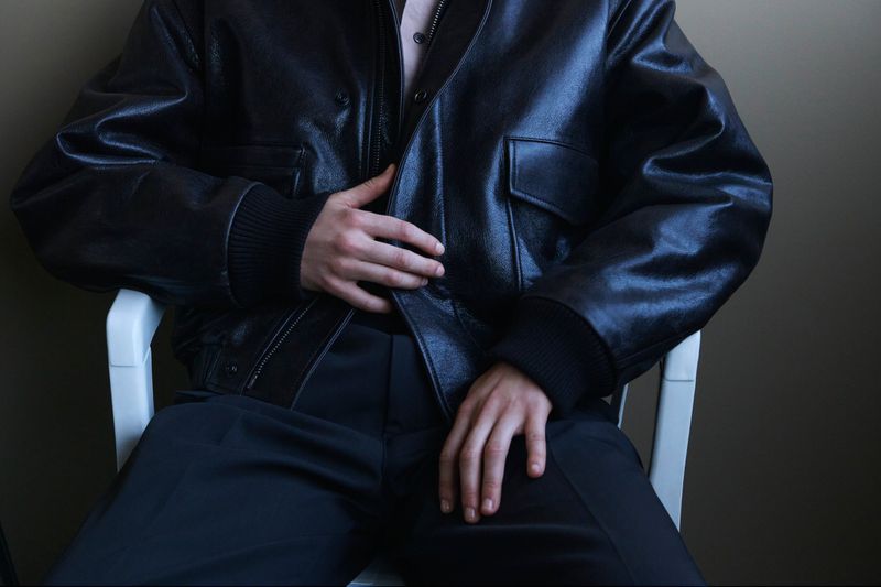 Close-up of a person sitting in a white chair wearing a glossy black leather bomber jacket and dark trousers for the Lemaire lookbook.