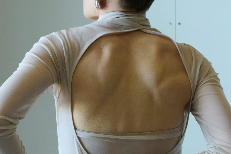 Close-up back view of a model wearing a sand beige long-sleeve top with a large circular cutout exposing the shoulder blades.