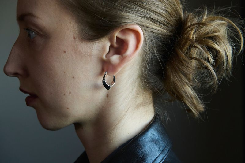 Woman in profile with hair in a bun wearing a curved silver hoop earring and a black leather jacket against a neutral background.