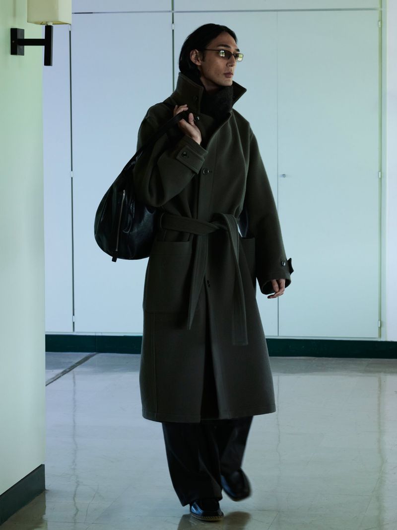 Model wearing a dark green belted coat and carrying a black shoulder bag walking indoors along a hallway