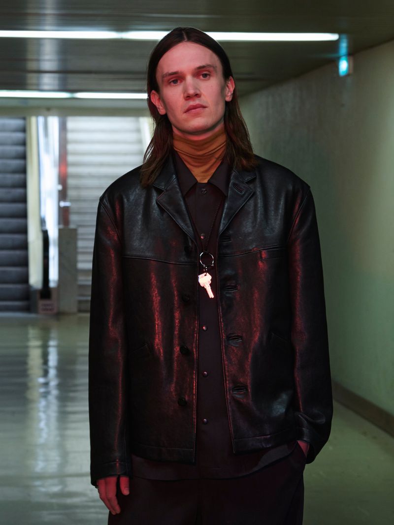 Model Moritz Thoma wears a black leather jacket over a camel turtleneck in a transit corridor for Lemaire.