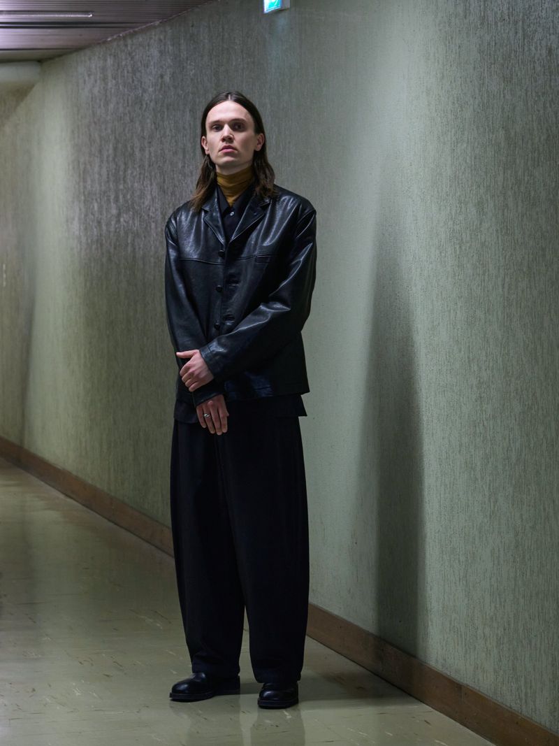 Model wearing a black leather buttoned jacket, yellow turtleneck, and wide-leg black trousers standing in an interior hallway.