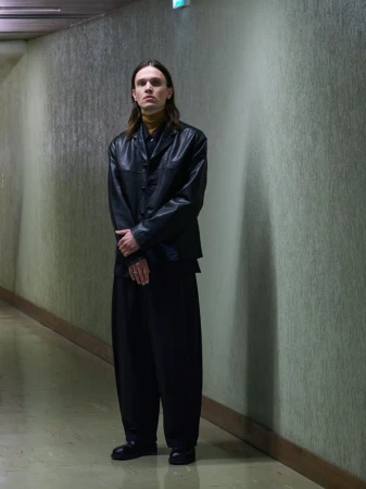 Model wearing a black leather buttoned jacket, yellow turtleneck, and wide-leg black trousers standing in an interior hallway.