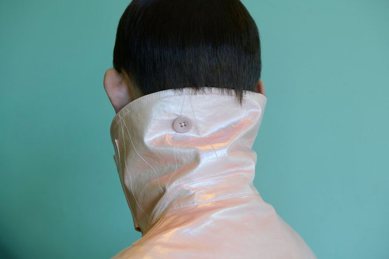 Back view of a person with short dark hair wearing a high-collared pale pink iridescent coat against a mint green background.