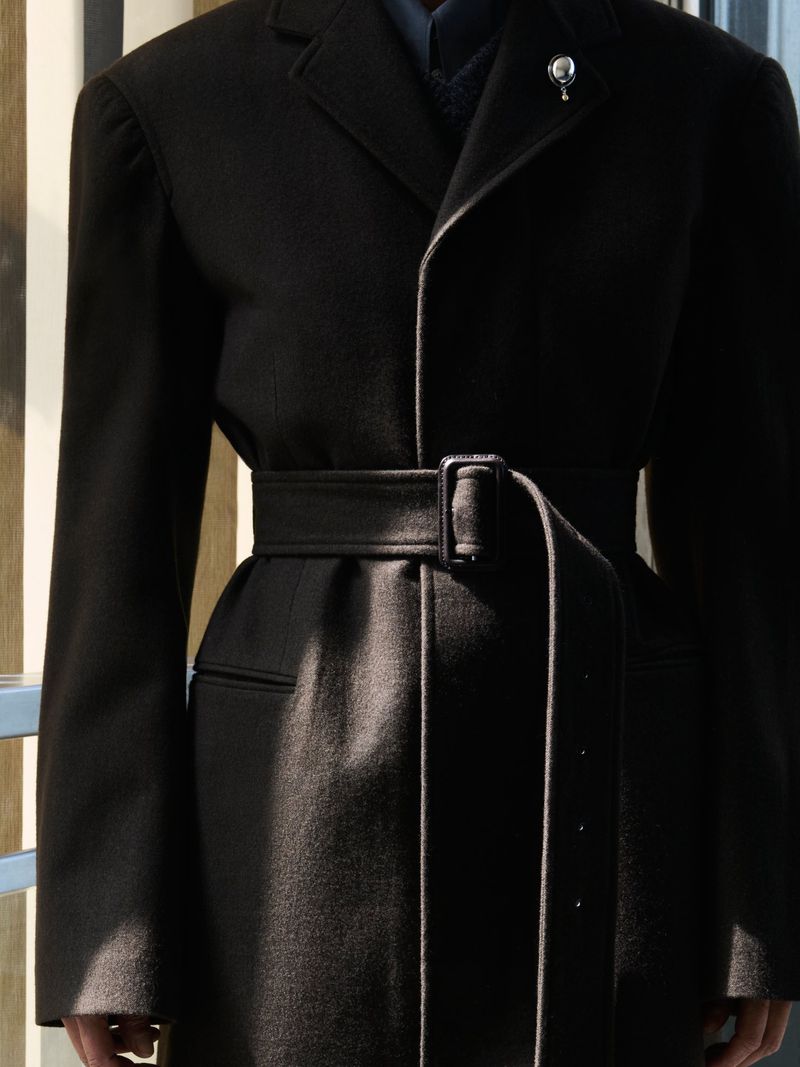 Close up of a tailored black belted coat with a lapel brooch cinched at the waist by a wide fabric belt.