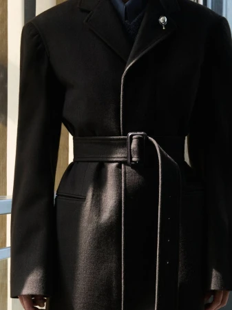 Close up of a tailored black belted coat with a lapel brooch cinched at the waist by a wide fabric belt.
