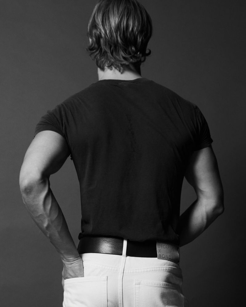 Model Brennan Aldred seen from behind wearing a black t-shirt and white jeans against a dark studio background.