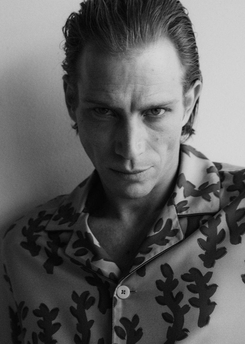 Monochromatic portrait of a fair-skinned adult male with slicked-back hair, wearing a patterned shirt and posing indoors.